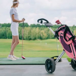 A person dressed in white golf attire holds a club while standing on a green, ready to make their shot. In the foreground, a pink and black golf bag is attached to a cart, subtly branded with payments logos.