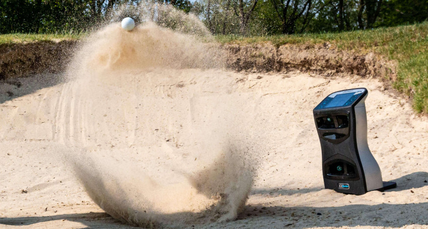 A golf launch monitor, monitored by a state-of-the-art golf launch monitor, hits a ball out of a sand bunker, causing a large spray of sand.