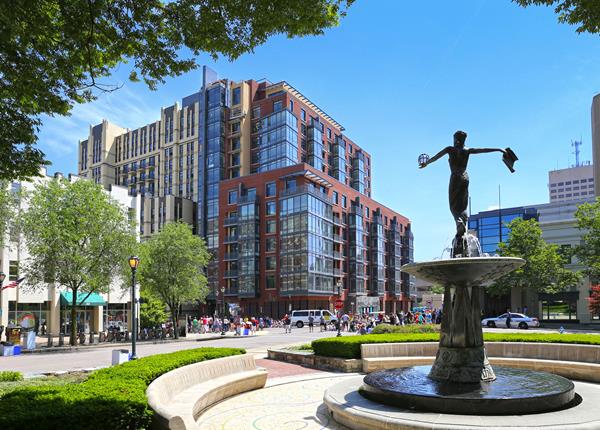 A modern cityscape in Rockville MD features a fountain surrounded by trees and benches. In the backdrop, towering buildings overlook a bustling street filled with people and cars, capturing the hustle while nearby, enthusiasts delve into custom golf area.