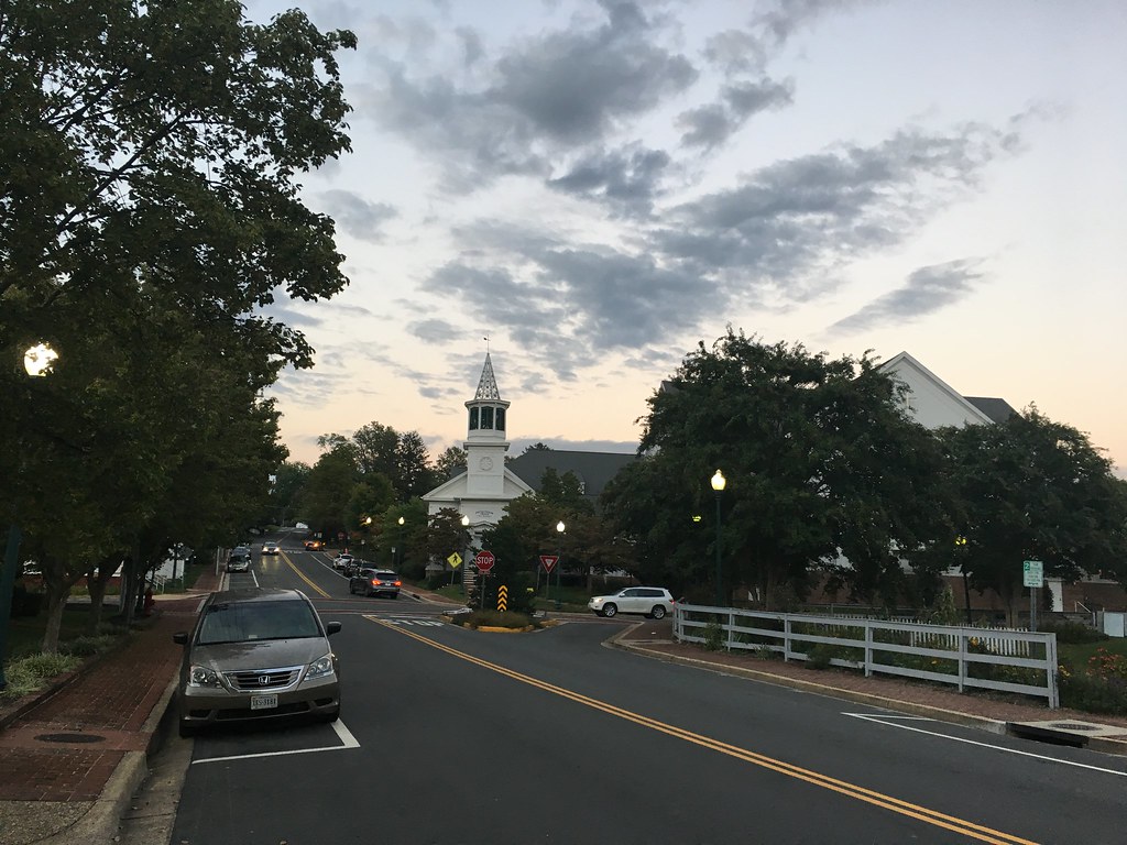 A small town street in Vienna, VA, features a charming white church with a steeple, surrounded by trees and parked cars, under a cloudy sky at dusk. Nearby, locals enjoy the art of custom golf club to perfect their game.