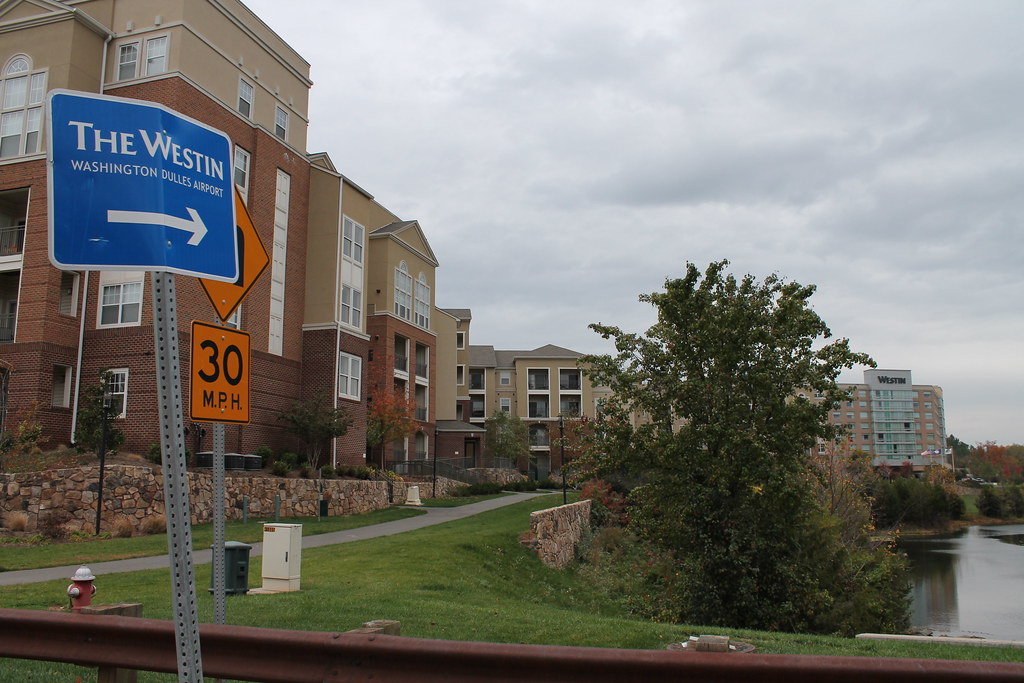 A sign directs to The Westin near a residential area with a 30 MPH speed limit, not far from where custom golf club fitting services are offered. A river runs alongside the pavement, and the sky is overcast.