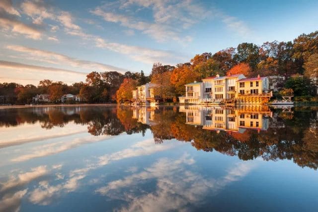 Houses by a tranquil lake with autumn trees and the gentle hum of a nearby golf club fitting in harmony with nature, all reflected under a partly cloudy sky.