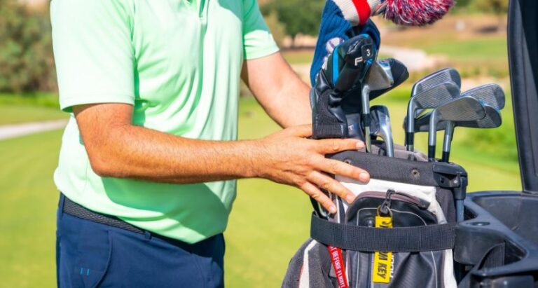 A person wearing a light green shirt and blue pants arranges golf clubs in a golf bag on a sunny golf course.