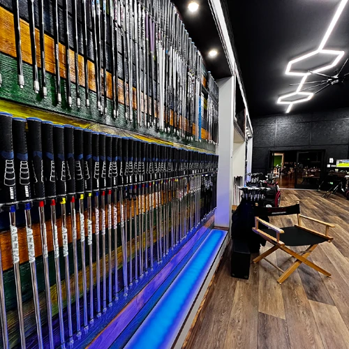 A wall display of golf clubs in a modern store with wood flooring, a director's chair, and illuminated blue shelf lighting.