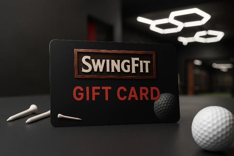 A SwingFit gift card is displayed upright on a table with two white golf tees and two golf balls nearby; the background is an indoor space with modern lighting.