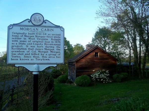 A historical marker for Morgan Cabin stands in front of a small wooden cabin surrounded by green grass, bushes, and trees under a blue sky.