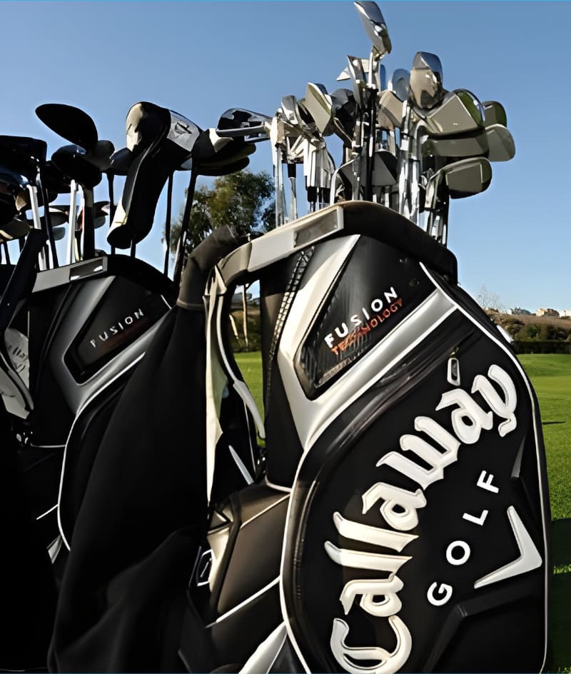 Close-up of Callaway golf bags filled with clubs standing on a grassy area, with trees and a clear sky in the background.