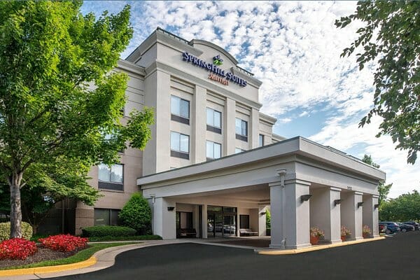 Four-story SpringHill Suites hotel building with a covered entrance, surrounded by green trees and landscaping, under a partly cloudy sky.