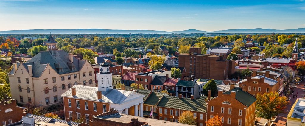 Aerial view of a small town with historic brick buildings, tree-lined streets, and distant mountains under a clear blue sky.