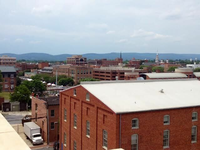 View of a cityscape with red brick buildings in the foreground, additional buildings and trees in the background, and distant hills under a cloudy sky.
