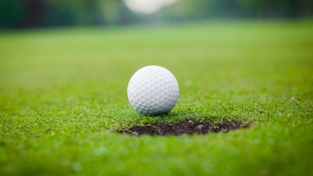 A white golf ball sits on the edge of a hole on a green golf course, with blurred grass and trees in the background.