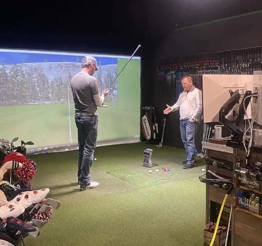 Two people stand on an indoor golf simulator; one holds a golf club while the other gestures, surrounded by golf equipment and digital screens.