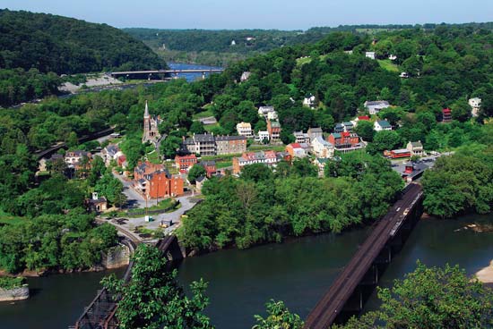 Aerial view of a small town with colorful buildings, a church, and bridges crossing a river, surrounded by green hills and trees.