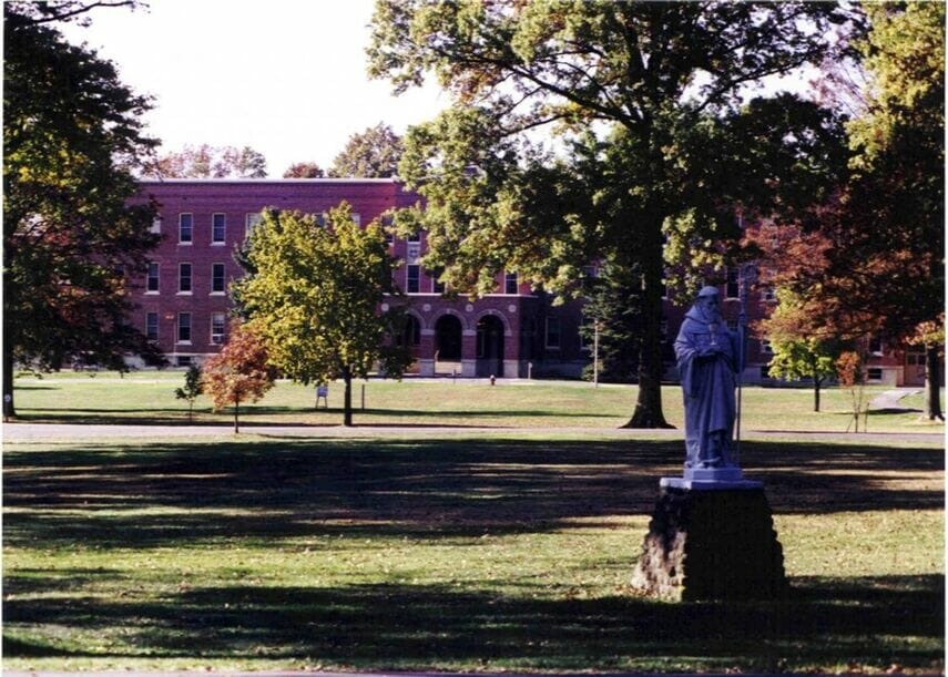 A brick building stands in the background behind a lawn with scattered trees and a statue on a pedestal in the foreground.