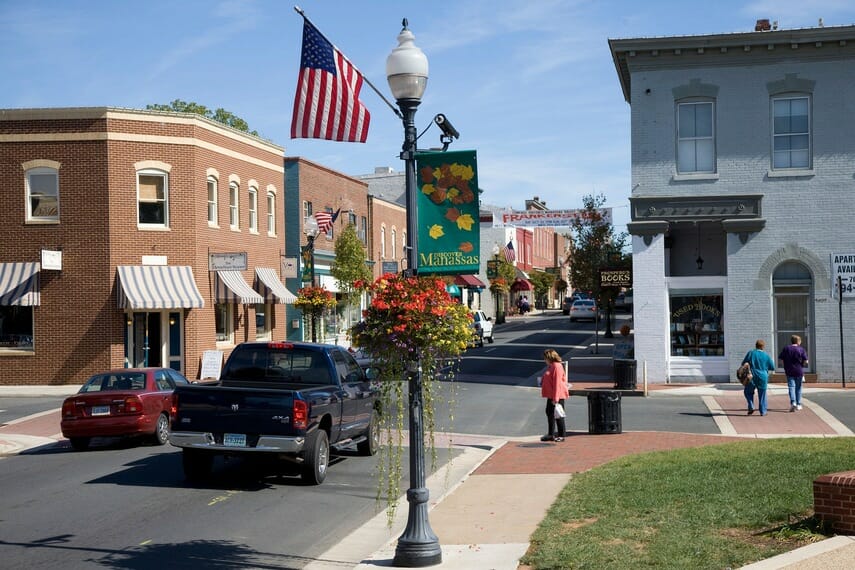 A small town street scene with brick buildings, cars, pedestrians, an American flag, and a hanging flower basket on a lamppost in Manassas, Virginia.