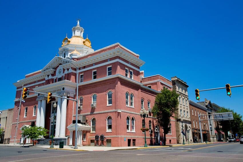 A large red-brick courthouse with white columns and a gold-domed cupola stands on a street corner under a clear blue sky, surrounded by historic buildings and traffic lights.