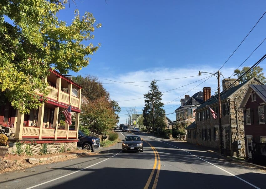 A car drives down a two-lane road lined with historic houses and trees under a clear blue sky in a small town.