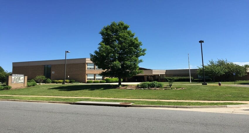 A two-story brick school building with a flat roof, surrounded by a lawn, a large tree, and a sign near the street under a clear blue sky.