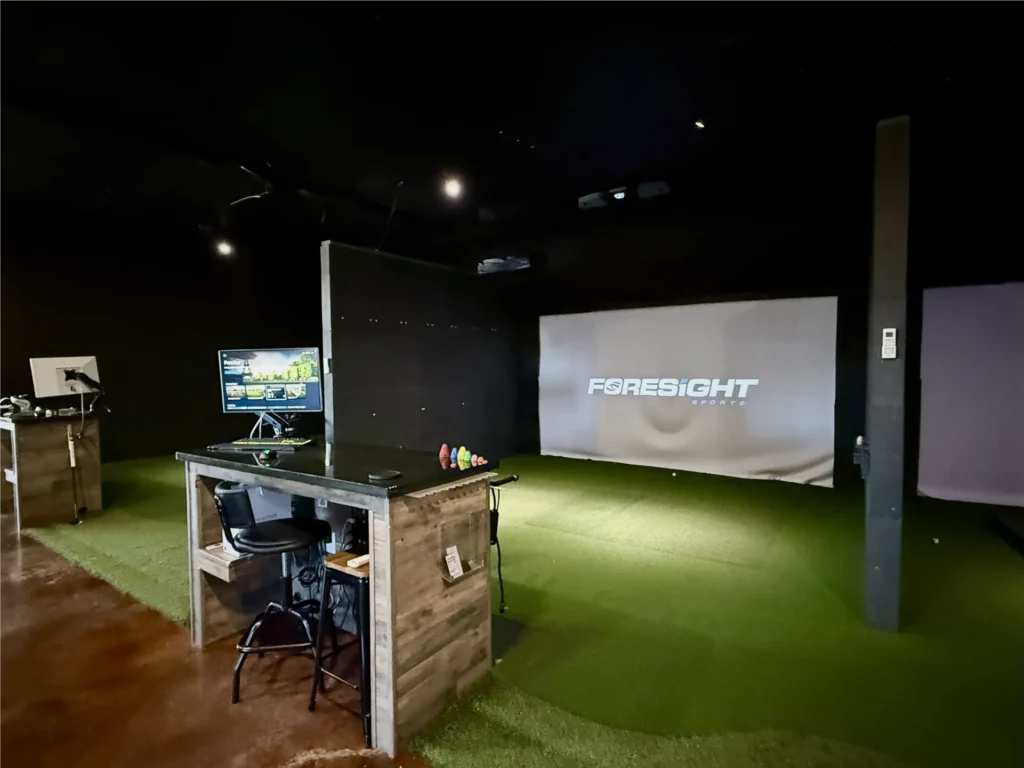 An indoor golf simulator setup with a desk, computer monitors, a chair, and a large FSRESiGHT screen on a turf floor in a dimly lit room.