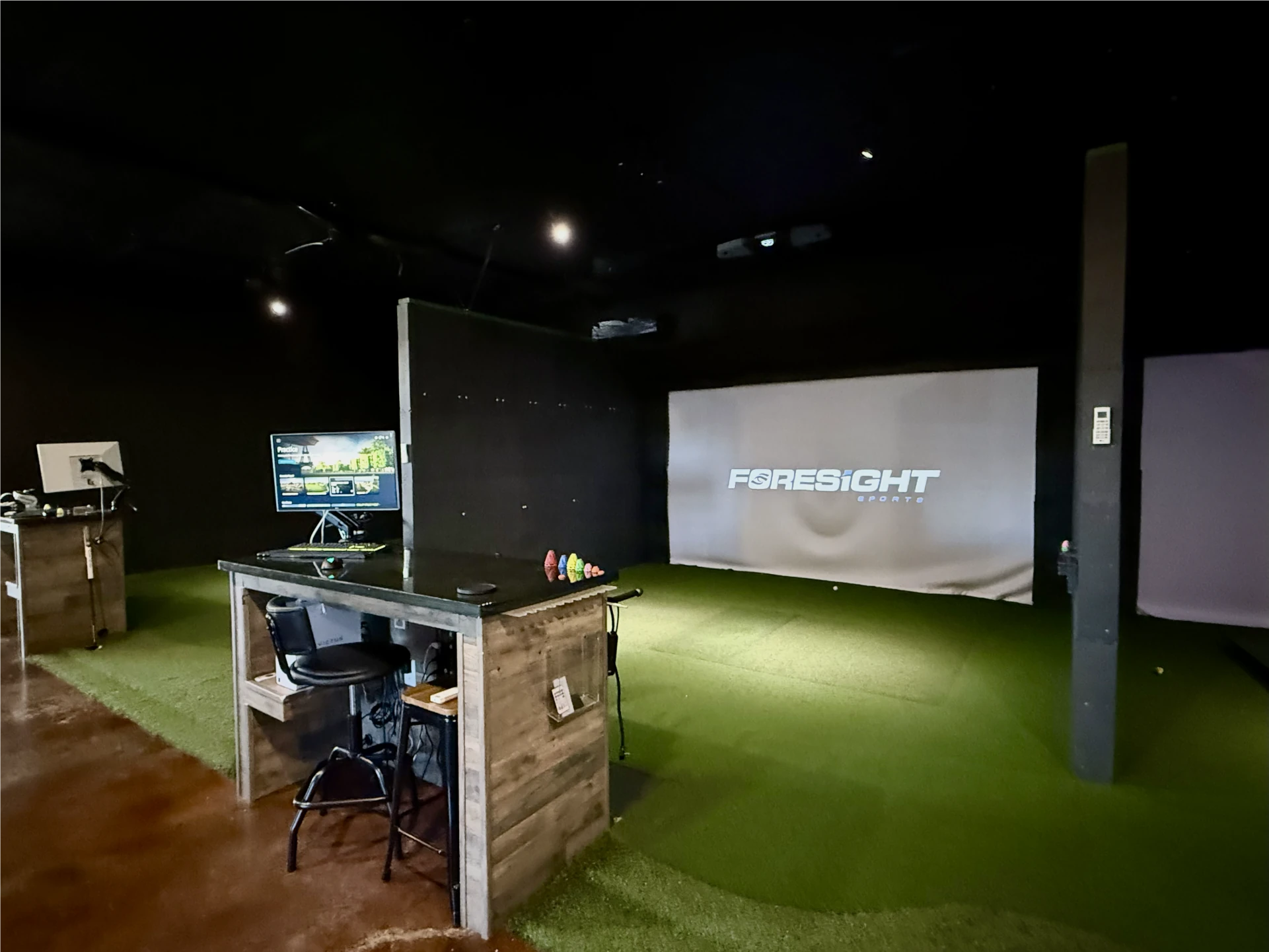 An indoor golf simulator setup with a desk, computer monitors, a chair, and a large FSRESiGHT screen on a turf floor in a dimly lit room.