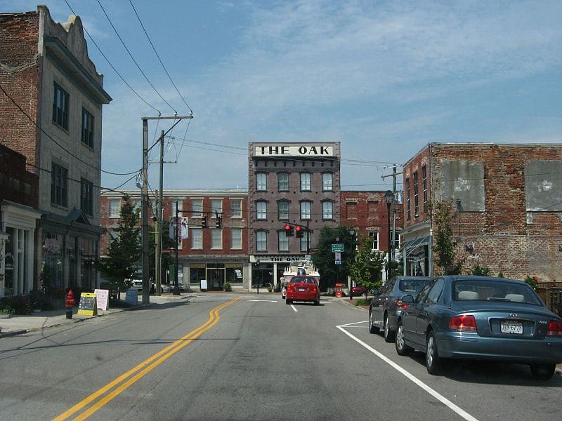 A small downtown street with parked cars, brick buildings, and a sign reading "The Oak" on a central building under a clear sky.