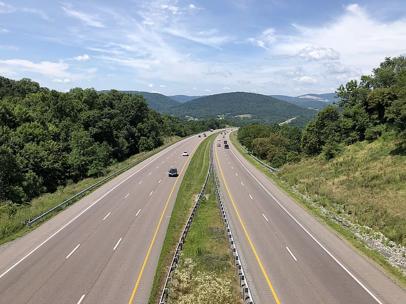 A divided highway with several cars traveling in both directions, surrounded by green hills and trees under a partly cloudy sky.