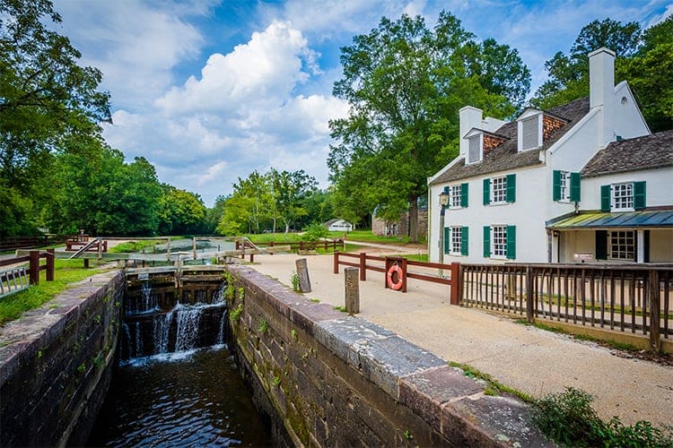 Historic canal lock with flowing water beside a white building with green shutters, surrounded by trees and a partly cloudy sky.
