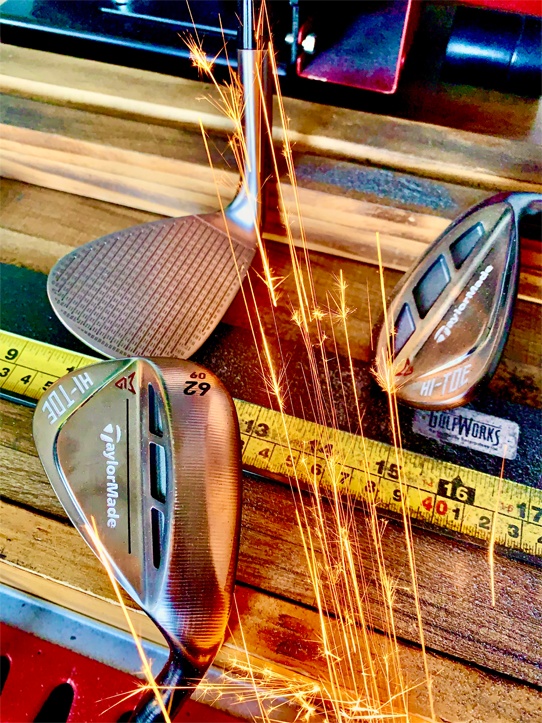 Three TaylorMade golf wedges on a workbench with a tape measure, as sparks fly nearby, possibly from grinding or cutting metal.