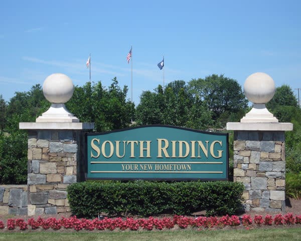 A stone entrance sign reads "South Riding Your New Hometown," with bushes, flowers, and flagpoles in the background.