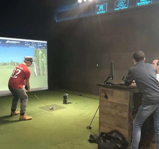 Three people are in a room with a golf simulator; one is preparing to swing a club while two others watch from behind a wooden desk with a computer.