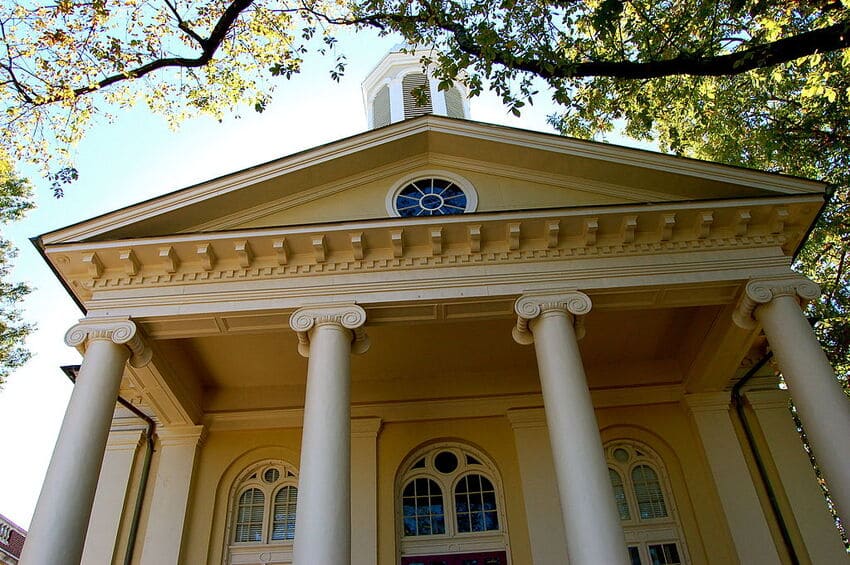 The image shows the entrance of a yellow neoclassical building with tall columns, arched windows, and a small cupola on the roof, partially shaded by tree branches.