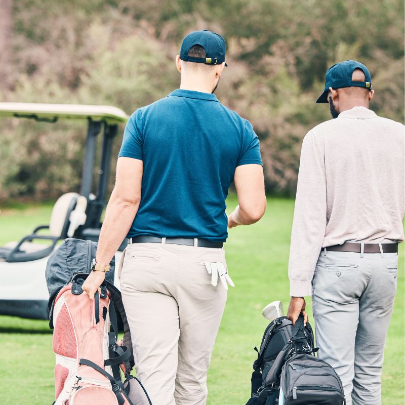 Two men wearing golf attire walk on grass carrying golf bags, with a golf cart nearby in the background.